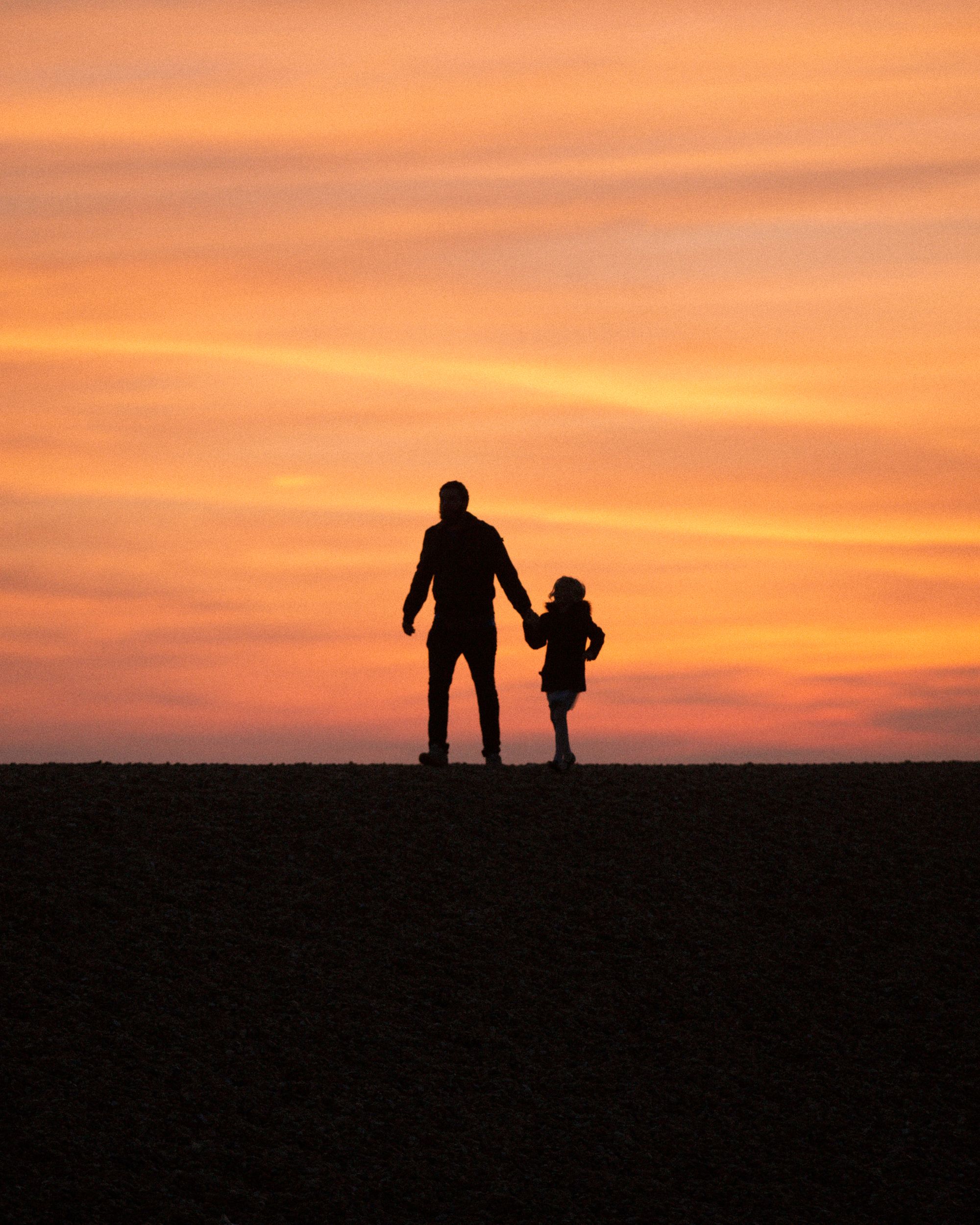 Silhouette eines Vaters, der bei Sonnenuntergang Hand in Hand mit seinem Kind auf einer Anhöhe steht